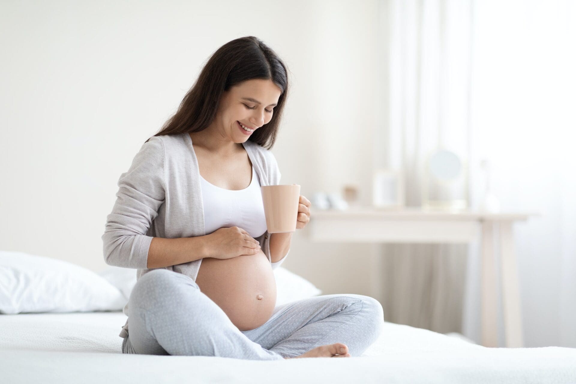 Smiling pregnant woman sitting on bed, drinking tea