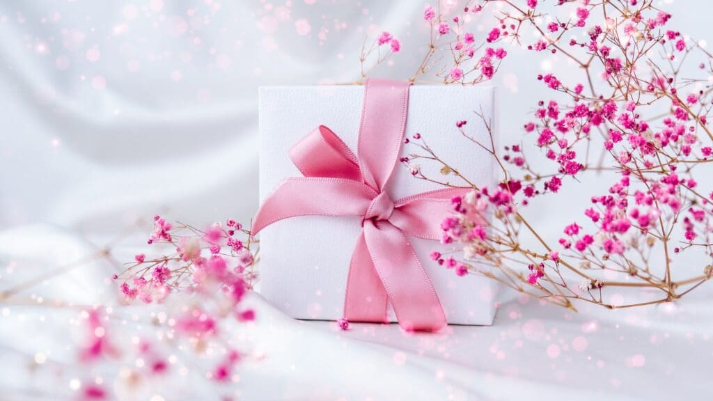 White present box with pink ribbon and small pink flowers on white silk fabric background. Greeting